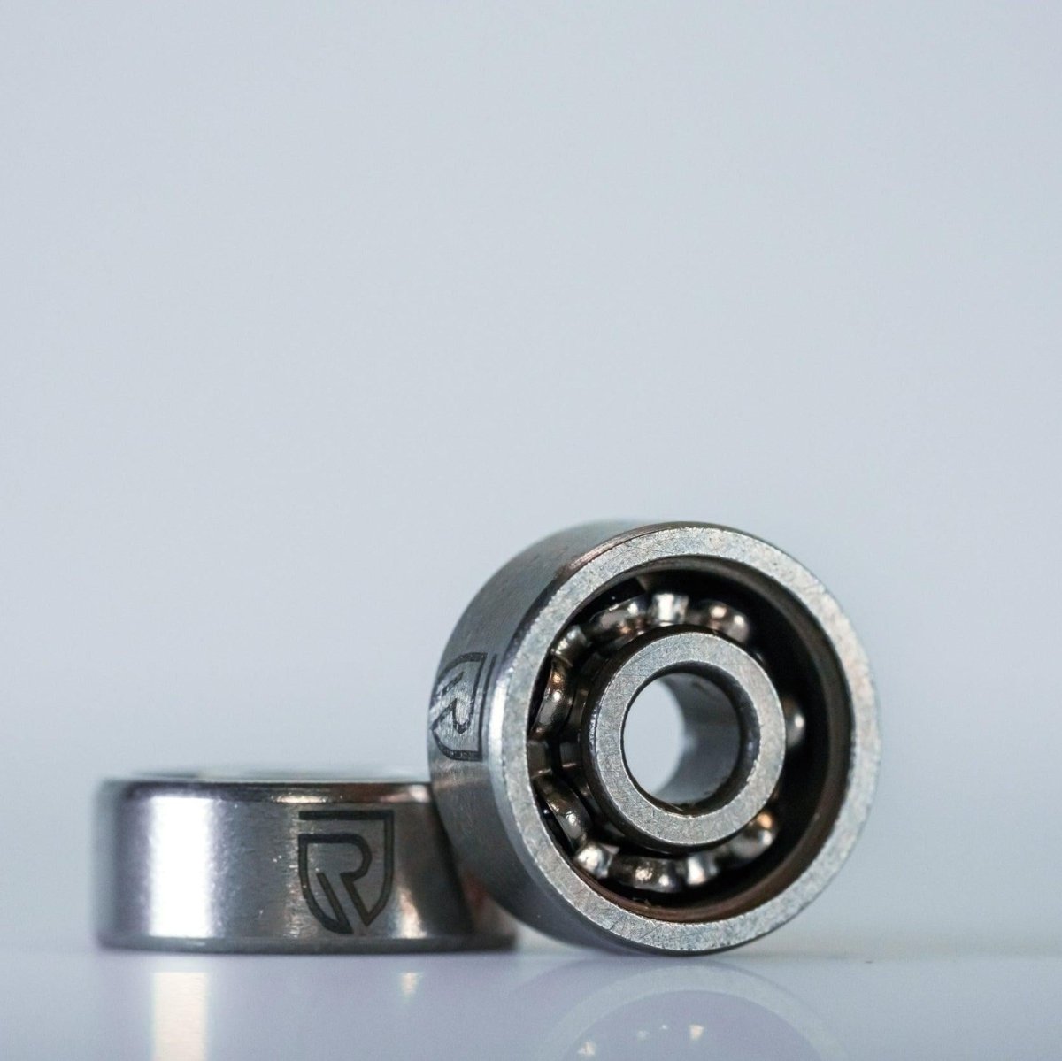 Abgebildet sind zwei RL-TNG Edelstahl Knob Kugellager 4x7x2,5 von RL-Angelrollentuning: Ein ABEC 5-Lager steht aufrecht und gibt den Blick auf die Kugeln frei, während ein anderes flach dahinter liegt, beide mit einem eingravierten Logo auf dem Außenring vor einem schlichten Hintergrund.