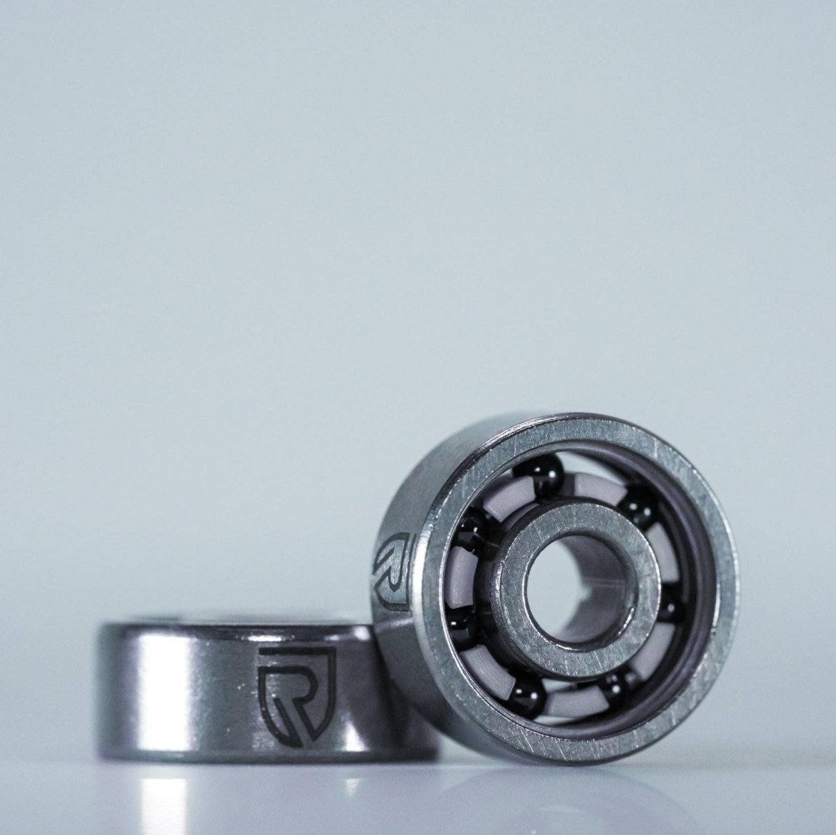 Zwei metallische RL-TNG High Speed Keramik Kugellager von RL-Angelrollentuning, gekennzeichnet mit dem stilisierten R-Logo, sind auf einem hellen Hintergrund abgebildet – eines aufrecht und gibt den Blick auf die Kugeln frei, eines flach dahinter. ABEC 9, alle Größen, für Baitcaster-Angelrollen.