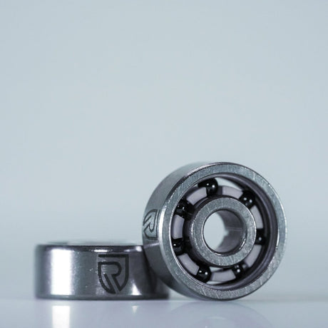 Zwei RL-TNG High Speed Keramik Spulenlager für Shimano SLX sind auf einer weißen Fläche abgebildet: eines aufrecht, so dass die inneren Kugeln zu sehen sind, und das andere liegend. Auf beiden Kugellagern ist seitlich das RL-TNG-Logo eingeätzt.