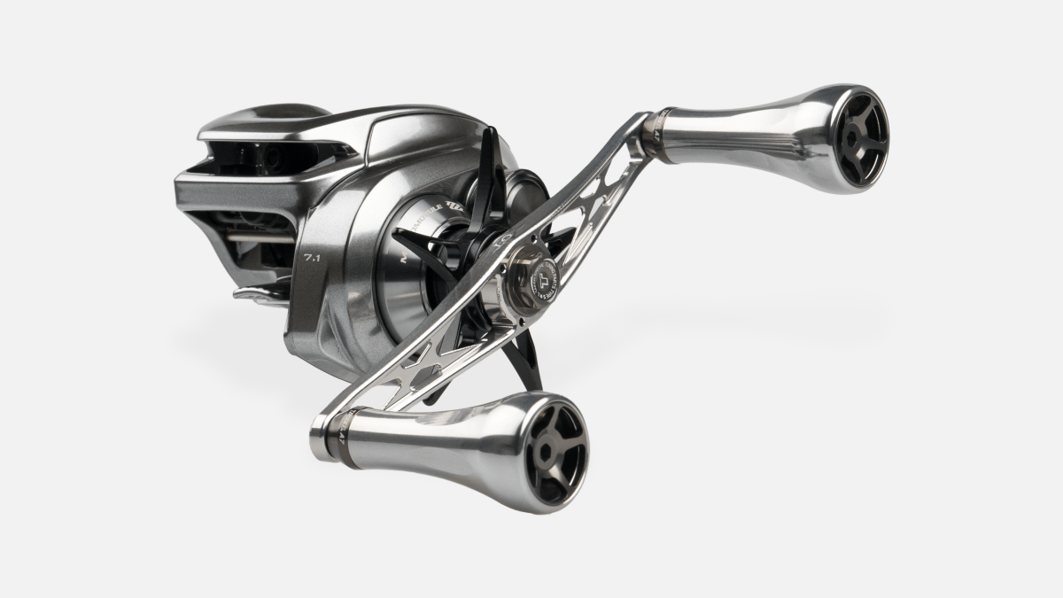 Der Taken GT106 Griff von Taken ist ein Griff aus Duraluminium mit A7 Aluminiumknöpfen für Shimano und Daiwa. Er ist in Silber mit einer großen Spule und einem Metallrahmen vor einem schlichten weißen Hintergrund dargestellt.