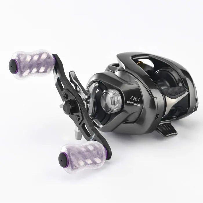 Auf weißem Hintergrund ist eine schwarze Baitcasting-Rolle mit zwei transparenten Gomexus 20-mm-Finesse-Touch-Knob-TPE-Griffen mit violetten Akzenten und einem Metallgehäuse mit verschiedenen Einstellrädern und Mechanismen abgebildet.
