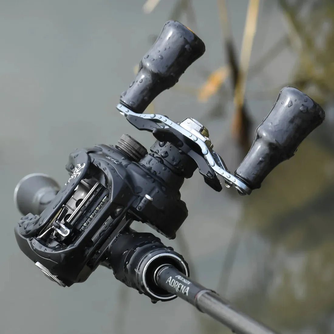 Eine Nahaufnahme einer schwarzen Angelrolle mit Wassertropfen, mit dem Gomexus Carbon Forged F24 Knob von Gomexus in ergonomischem Design, befestigt an einer über Wasser gehaltenen Rute mit verschwommenem Schilf im Hintergrund.