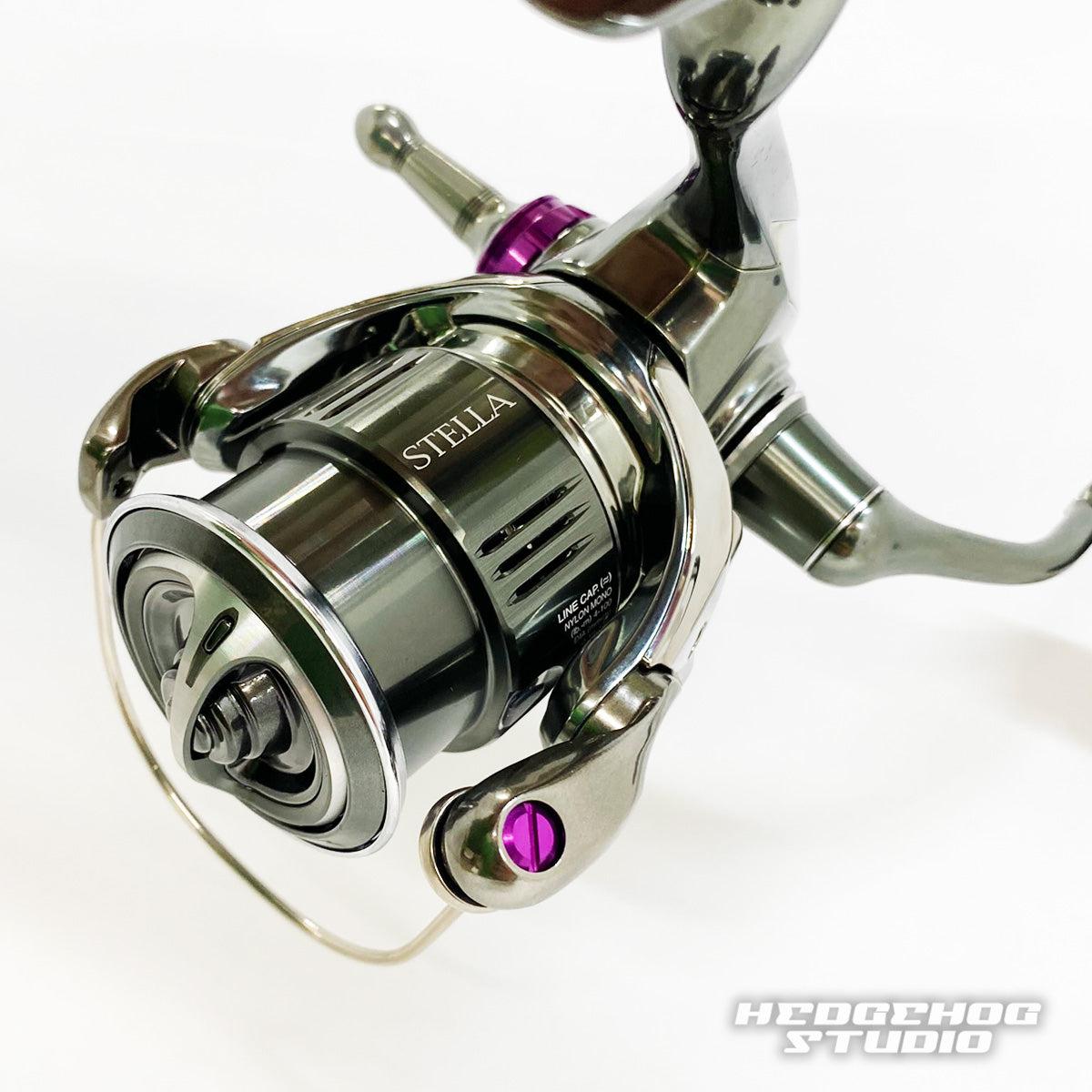 Ein Nahaufnahmefoto zeigt eine Shimano Stella-Rolle mit detaillierten Teilen, violetten Akzenten und einem Hedgehog Studio Extra Super Duralumin Schraube für Schnurlaufröllchen-Upgrade mit den Logos STELLA und HEDGEHOG STUDIO auf der Rolle.