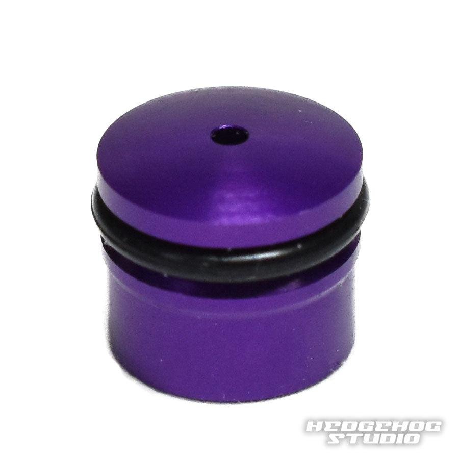 Eine kleine, zylindrische, violette Duraluminium-Knopfkappe für Daiwa Zaion mit schwarzem O-Ring oben und Loch in der Mitte. Das Bild befindet sich auf weißem Hintergrund mit der Aufschrift „HEDGEHOG STUDIO“ in der unteren rechten Ecke.