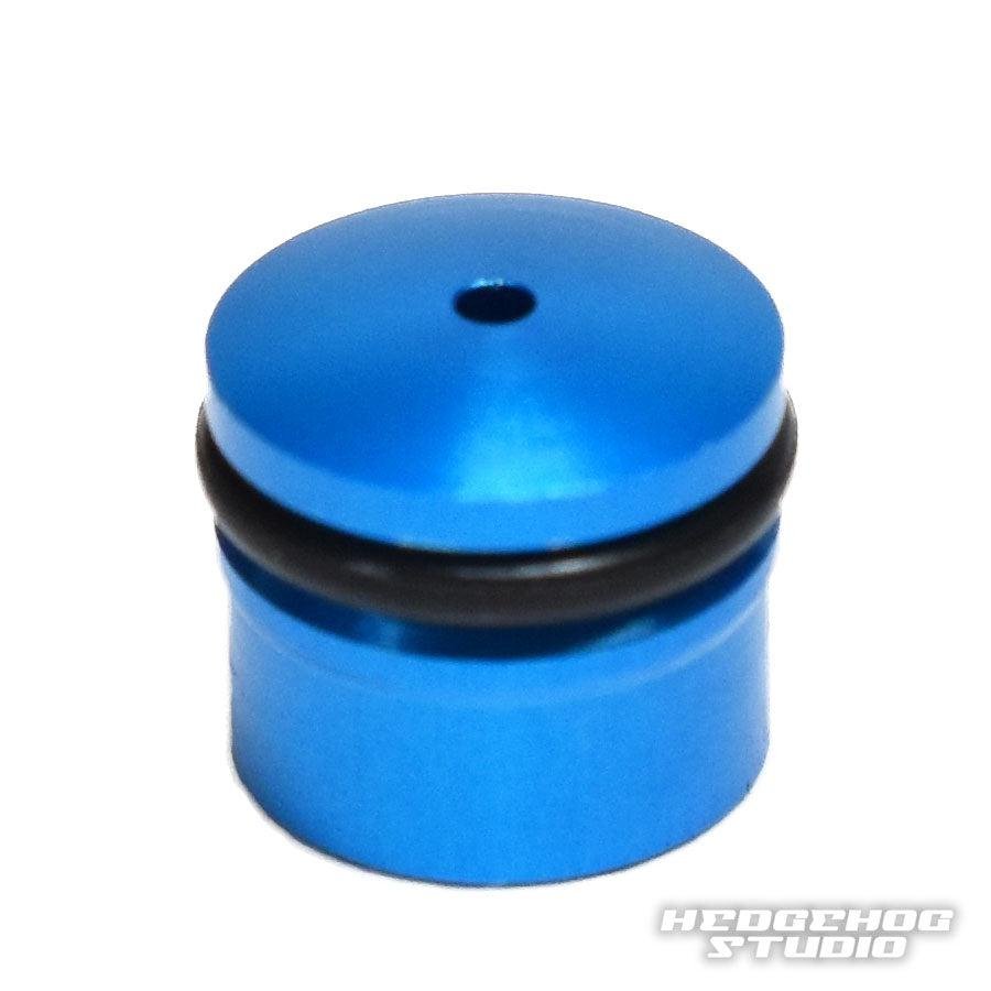 Eine kleine, zylindrische blaue Hedgehog Studio-Knopfkappe für Daiwa Zaion aus Duralumin mit abgerundeter Oberseite und einem schwarzen O-Ring in der Mitte ist auf einem weißen Hintergrund mit dem Hedgehog Studio-Logo in der unteren rechten Ecke abgebildet.