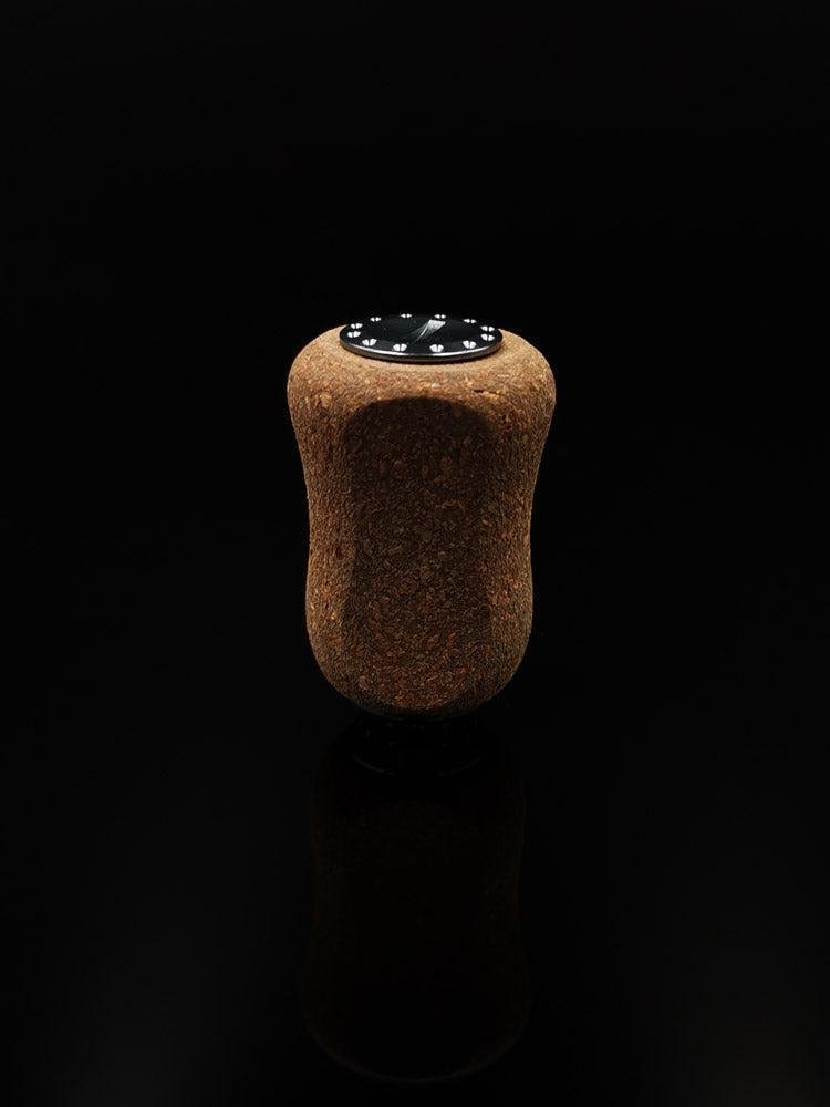 Ein einzelner RL-TNG Kork Knob 20 mm | Griff | Universal Fit von RL-Angelrollentuning mit dunkler Oberseite und Drahtmarkierungen steht aufrecht auf einer glatten schwarzen Oberfläche vor einem schlichten schwarzen Hintergrund.
