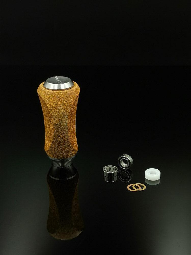 Ein RL-TNG Kork Knob von RL-Angelrollentuning mit strukturierter zylindrischer Form und mehrfarbigem Design steht aufrecht auf einer schwarzen Oberfläche neben einzelnen Unterlegscheiben, Lagern und kleinen Metall-/Kunststoffteilen einer Angelrolle.