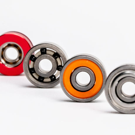 Vier Kugellager von Shimano Kugellager by RL-TNG sind diagonal auf weißem Hintergrund angeordnet, jedes mit einer anderen Ringfarbe - rot, schwarz, orange und einfarbiges Metall - und zeigen so deutlich ihr High-Speed-Design und ihre feinen Details.