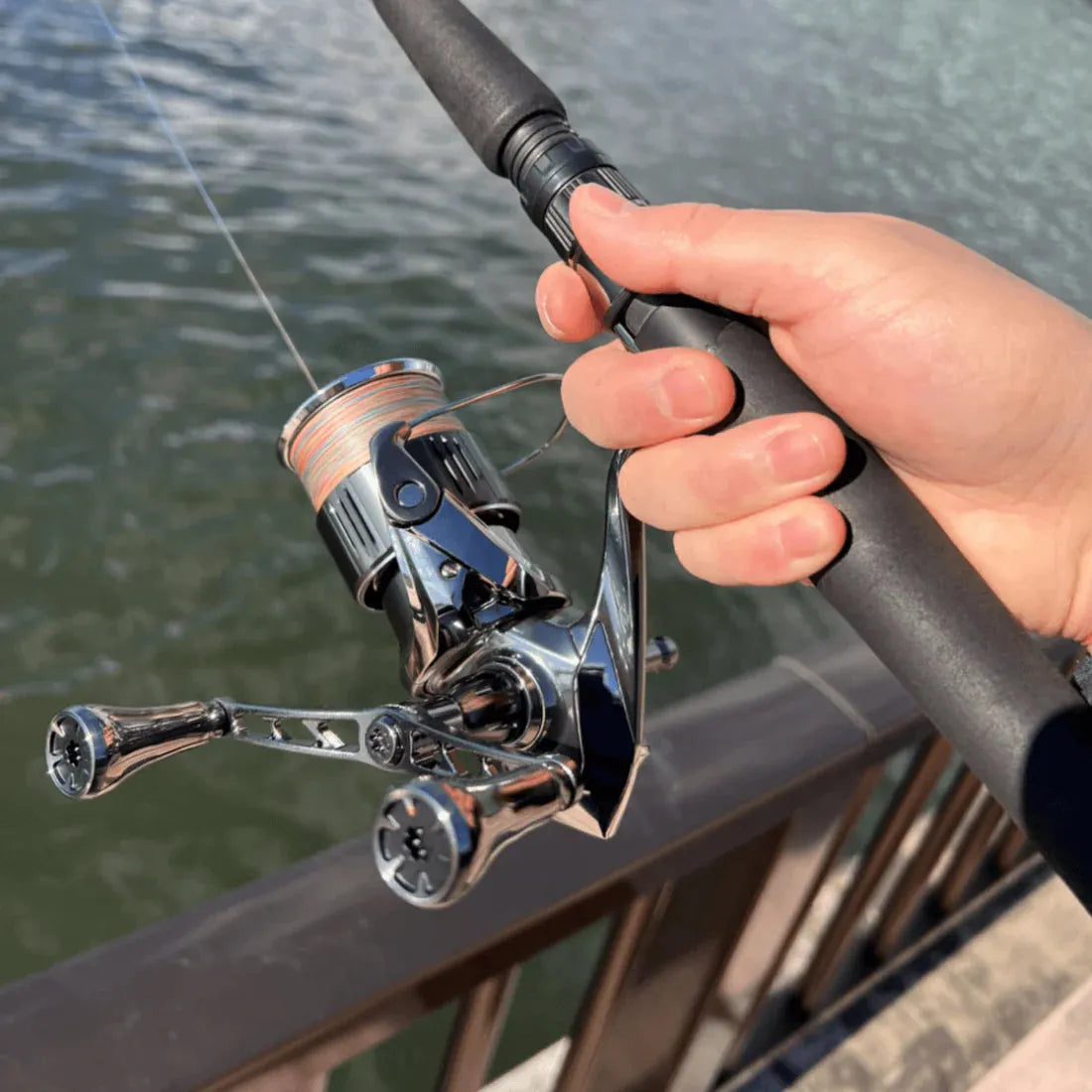 Eine Hand hält eine Angelrute mit einem Gomexus-Doppelgriff 98–82 mm mit T20-Knöpfen für Shimano, positioniert über dem Wasser neben einem Metallgeländer.