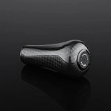 Ein schwarzer, strukturierter Gomexus FA30 Carbon-Knopf von Gomexus aus hochwertigem Toray-Carbon sitzt auf einer glänzend schwarzen Oberfläche. Weiße Markierungen und Zahlen auf der Oberseite weisen auf die Kompatibilität mit 5-Gang-Schaltgetrieben hin.