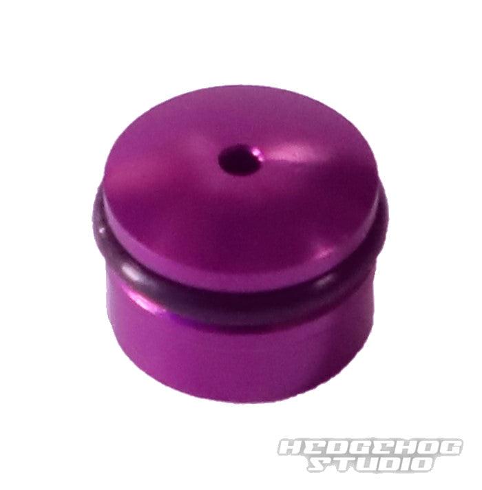 Eine kleine, zylindrische, metallisch-lila Hedgehog Studio-Knopfkappe für Daiwa Finesse-Knöpfe mit einem zentralen oberen Loch und einem schwarzen O-Ring, abgebildet auf einem weißen Hintergrund mit einem schwachen HEDGEHOG STUDIO-Wasserzeichen.