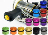 Ein Hedgehog Studio Knob Cap | Daiwa Finesse Knob ist neben einer Angelrolle mit schwarzem Griff abgebildet, umgeben von zwölf metallisch gefärbten Duraluminiumknöpfen in Blau, Lila, Rot, Grün, Gold, Silber und Schwarz.