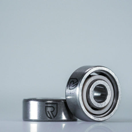 Zwei RL-TNG High Speed Keramik Kugellager (ABEC 9, Daiwa, 5x11x4 & 3x10x4) von RL-Angelrollentuning liegen auf einer glatten Oberfläche mit stilisierten R-Logos. Perfekt für Ihre Angelrolle. Heller, aufgeräumter Hintergrund.