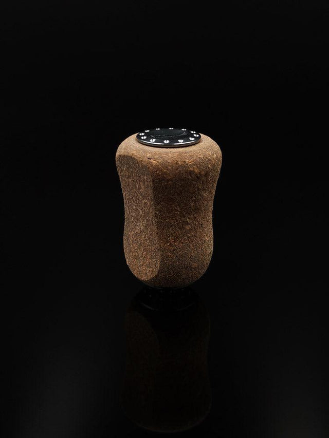 Der RL-TNG Kork Knob 20mm von RL-Angelrollentuning – ein einzelner Korkgriff mit schwarzer Folienoberfläche – steht aufrecht vor einem schwarzen Hintergrund und erzeugt eine subtile Reflexion darunter. Universelle Passform für vielseitigen Einsatz.