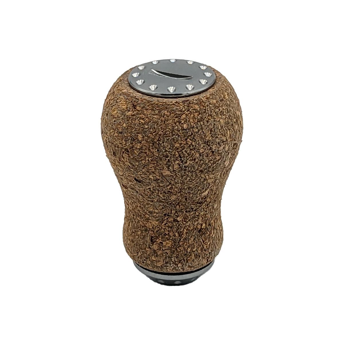 Der RL-Angelrollentuning RL-TNG Korkknopf 20 mm „Peanut“ Universal Fit verfügt über eine abgerundete Korkabdeckung, eine metallische Oberseite mit Schichtmuster und dekorative Nieten, inspiriert von Edelstahlkugellagern, isoliert auf Weiß dargestellt.