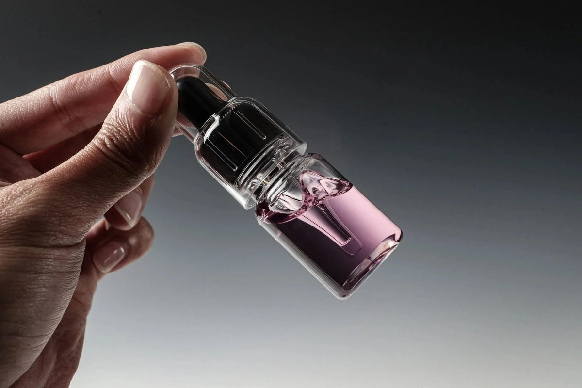 Eine Hand hält eine kleine Tropfflasche mit Glitch Oil Rosa von Glitch Oil Japan, gefüllt mit einer klaren, violett getönten Flüssigkeit, vor einem grauen Hintergrund mit Farbverlauf. Ideal zur Reduzierung von Korrosion und Verschleiß, insbesondere beim Salzwasserfischen.