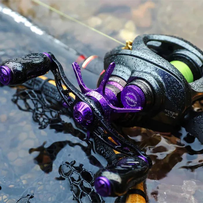 Eine Nahaufnahme einer Gomexus Aluminium-Spulenspannungsknopfkappe in Lila und Schwarz, die von Wassertropfen glitzert, während sie auf einer Angelrute ruht, die teilweise in seichtes Wasser eingetaucht ist.