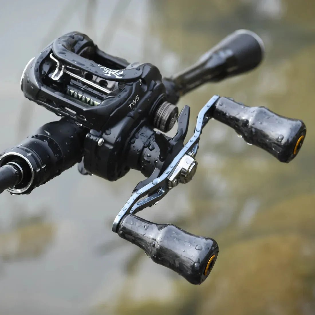 Eine schwarze Baitcastingrolle mit Wassertropfen, ausgestattet mit einem Gomexus Carbon Forged F24 Knob für ergonomische Handhabung, ist auf einer Stange über Wasser montiert.