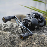 Der Gomexus Carbon Forged F24 Knob von Gomexus mit zwei Griffen und ergonomischem Design ist auf einer schwarzen Baitcasting-Angelrolle zu sehen, die auf einem großen Felsen im Freien ruht, mit verschwommenem Wasser und Schilf im Hintergrund.