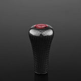 Ein Gomexus FA30 Carbon-Knopf aus Toray-Carbon mit strukturiertem Muster und roter runder Oberseite steht aufrecht auf einer reflektierenden schwarzen Oberfläche.