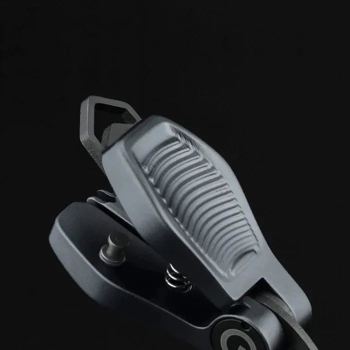 Eine Nahaufnahme des Gomexus SharkBite Schnurknipser Combo, ein graues, strukturiertes Metall-Klappwerkzeug mit geriffeltem Griff, Scharnier und Federmechanismus von Gomexus, vor einem schlichten schwarzen Hintergrund.