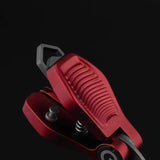 Eine Nahaufnahme des rot-metallischen Gomexus SharkBite Schnurknipser Combo mit strukturiertem Griff und freiliegender Feder, mit einem dunklen geometrischen Aufsatz, vor einem schwarzen Hintergrund.