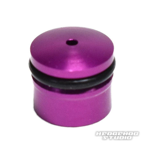 Eine kleine, zylindrische, metallisch-lila Hedgehog Studio-Knopfkappe für Daiwa Zaion-Rollen mit abgerundeter Oberseite, schwarzem Gummiring in der Mitte und einem oberen Loch auf weißem Hintergrund mit dem Text HEDGEHOG STUDIO in der Ecke.