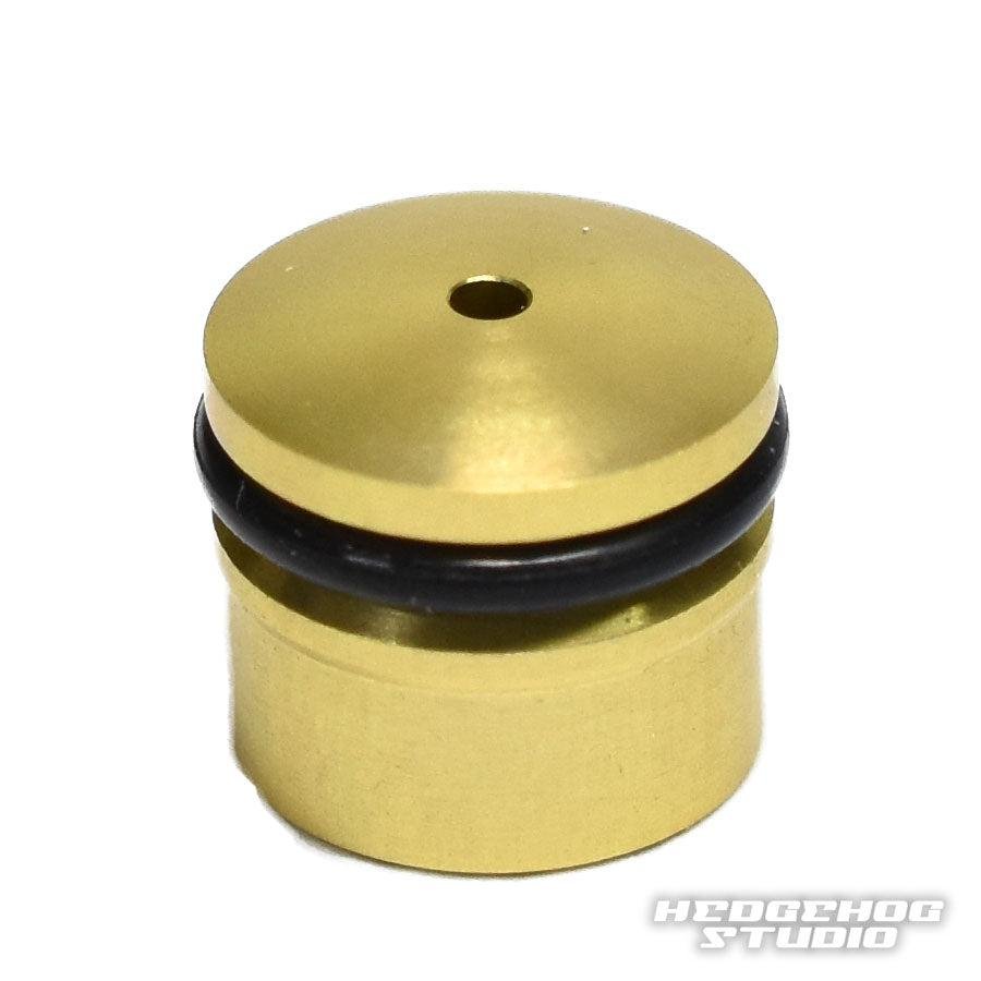 Eine kleine, zylindrische, goldene Hedgehog Studio Knob Cap für Daiwa Zaion mit zentralem Loch und schwarzem O-Ring am oberen Rand. Das HEDGEHOG STUDIO-Wasserzeichen erscheint in der unteren rechten Ecke des Bildes.