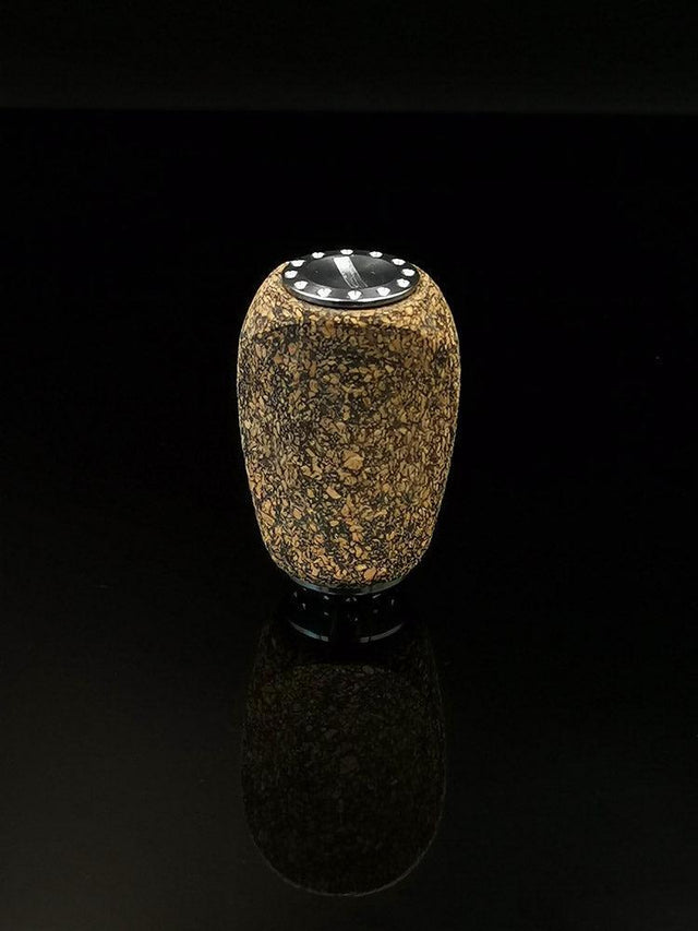 Der RL-TNG Korkknopf 20 mm | Schlank | Universal Fit von RL-Angelrollentuning ist ein kleiner, zylindrischer Knopf mit strukturiertem Korkkörper, schwarzer Oberseite und metallischen Edelstahlkugellager-Akzenten, der auf einer glänzend schwarzen Oberfläche vor dunklem Hintergrund steht.