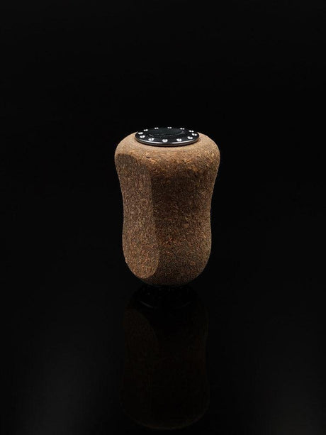 Der RL-TNG Kork Knob 20mm von RL-Angelrollentuning – ein einzelner Korkgriff mit schwarzer Folienoberfläche – steht aufrecht vor einem schwarzen Hintergrund und erzeugt eine subtile Reflexion darunter. Universelle Passform für vielseitigen Einsatz.