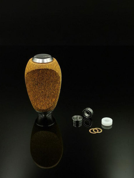 Ein RL-TNG Kork Knob von RL-Angelrollentuning mit strukturiertem braun-schwarzem Design steht aufrecht auf einer reflektierenden Oberfläche, daneben sind Metall- und Kunststoffkomponenten vor einem schwarzen Hintergrund angeordnet.