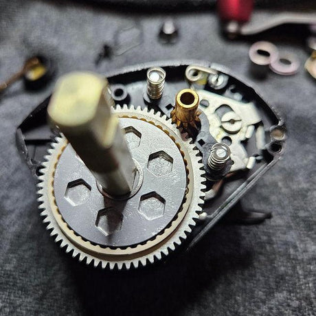 Nahaufnahme des Dragclickers für Daiwa Tatula 200 von Rollenwerk. Freiliegende Zahnräder und Komponenten mit Schwerpunkt auf dem Dragclicker-Mechanismus sind zu sehen; vor einem grauen strukturierten Hintergrund mit sichtbaren Kleinteilen.